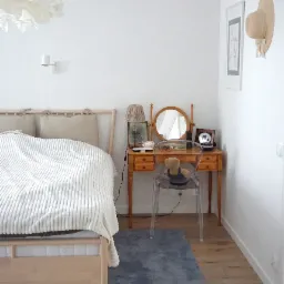 Jasna sypialnia w minimalistycznym stylu z drewnianym łóżkiem, toaletką z okrągłym lustrem i przezroczystym krzesłem, uzupełniona o dekoracje takie jak lampka, obrazek i kapelusz na ścianie.