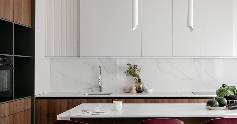 Nowoczesna kuchnia z białymi szafkami, marmurowym blatem i backsplashiem, oświetleniem tubowym oraz elementami drewnopodobnymi, z wyspą, na której leży otwarta książka i stoi biały kubek.