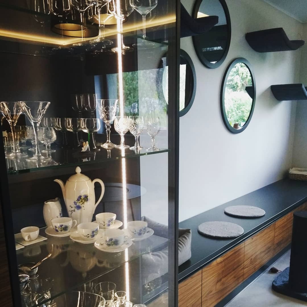 Szklana witryna z oświetleniem LED, zawierająca kolekcję kieliszków i porcelanowy serwis do herbaty w aranżacji wnętrza z okrągłymi lustrami i półkami dla kota na ścianie.