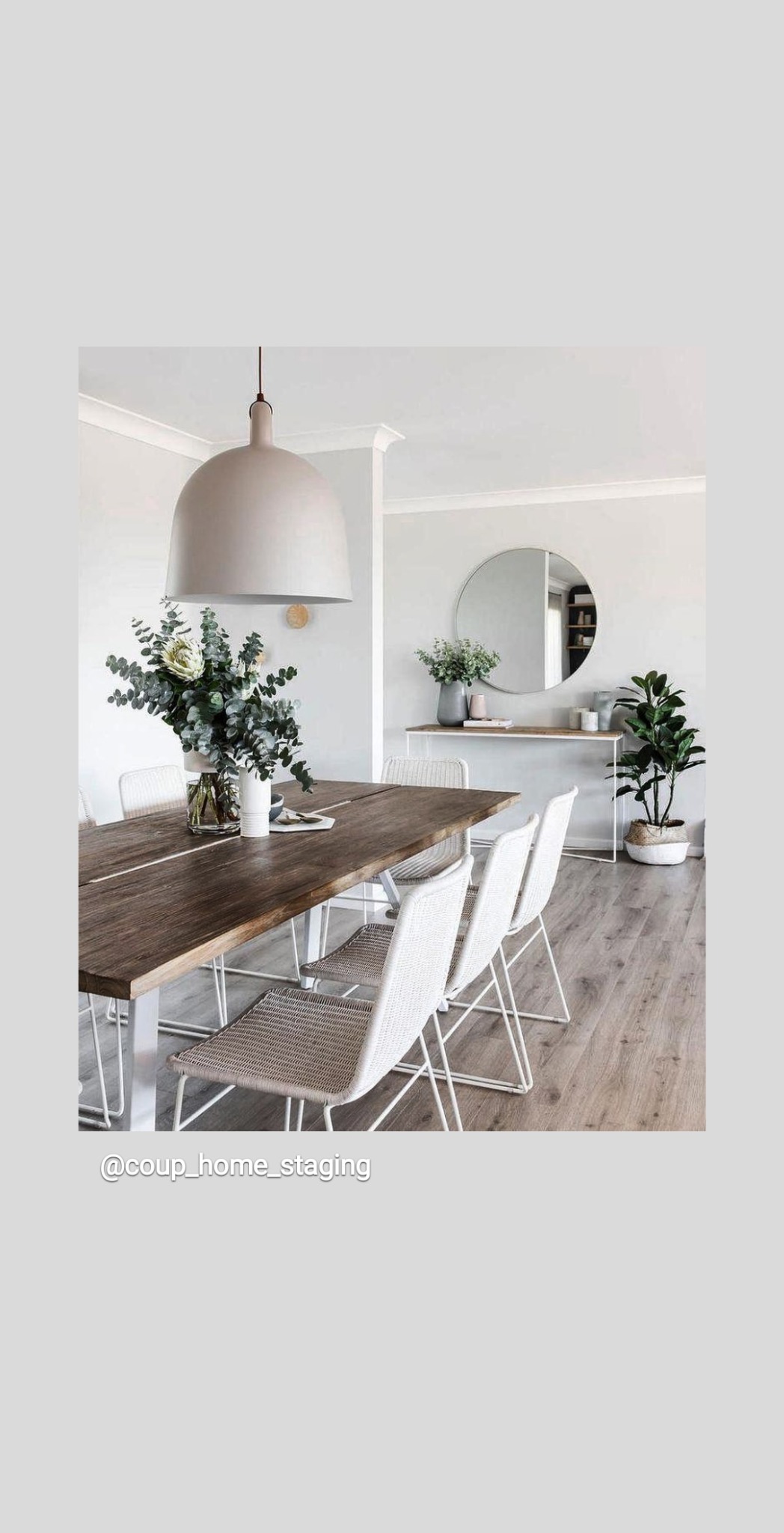 Jasne wnętrze jadalni z drewnianym stołem, białymi krzesłami z plecionym siedziskiem, okrągłym lustrem na ścianie i roślinami w wazonie oraz doniczce.