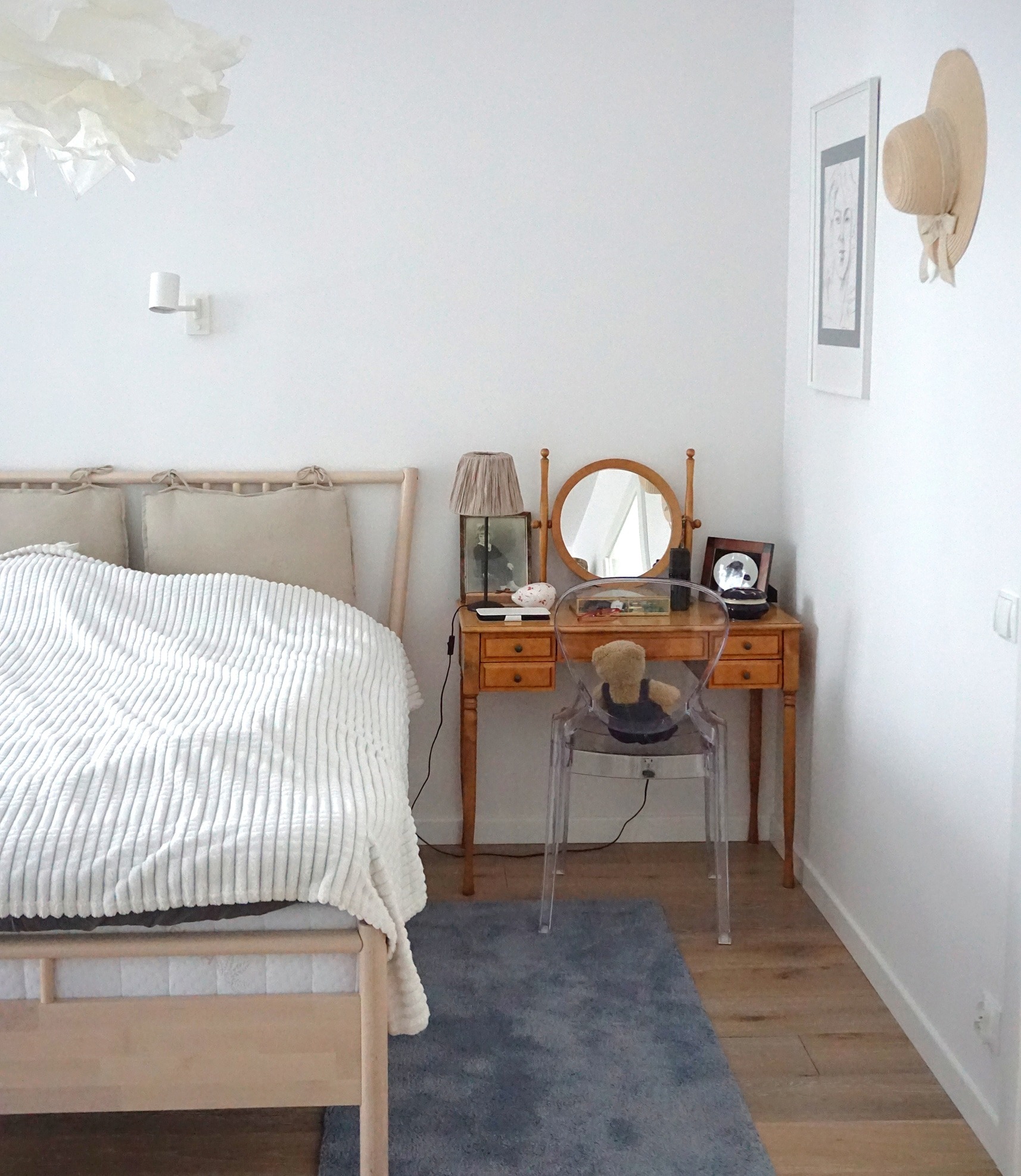 Jasna sypialnia w minimalistycznym stylu z drewnianym łóżkiem, toaletką z okrągłym lustrem i przezroczystym krzesłem, uzupełniona o dekoracje takie jak lampka, obrazek i kapelusz na ścianie.