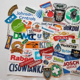 Kolekcja różnorodnych, haftowanych naszywek z logo firm i organizacji, ułożonych na białym tle, prezentująca szeroki zakres wzorów i kolorów, w tym logo marek spożywczych, motoryzacyjnych...
