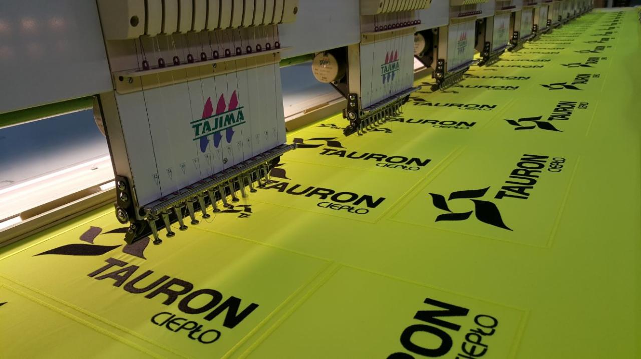 Wielogłowicowa maszyna hafciarska Tajima w trakcie haftowania czarnego logo Tauron Ciepło na jasnożółtym materiale.