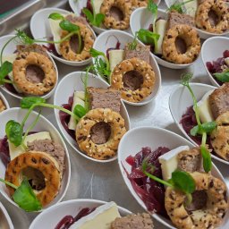 Global Chefs Mariusz Powierża - Apetizer w białej łódce: pasztet, ser brie, cebula i mini-bajgiel z ziarnami, udekorowany zielonym pędem. Elegancki finger food na srebrnej tacy.