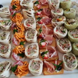 Dekoracyjna taca z różnorodnymi przekąskami: roladki z tortilli z widocznym farszem, kanapeczki z pastą, krakersy z szynką parmeńską i truskawkami, oraz piankowe bezy.