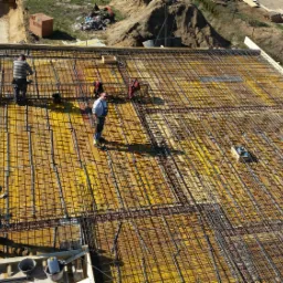 Zbrojenie płyty fundamentowej z żółtym szalunkiem, widok z góry, pracownicy w trakcie montażu.