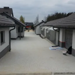 Nowoczesne domy murowane z szarymi dachami i jasnymi elewacjami, widok na wybrukowany podjazd pomiędzy budynkami, z elementami wykończeniowymi w trakcie realizacji.