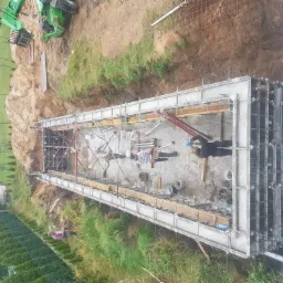 Budowa fundamentów pod budynek: szalunek z metalowych paneli, zbrojenie stalowe, widoczni pracownicy budowlani i sprzęt budowlany na placu budowy.