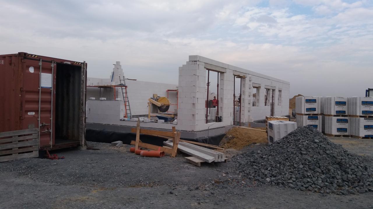 Wznoszenie ścian parterowego domu z białych bloczków, widoczny kontener budowlany, stosy bloczków i gruzu, oraz piła do cięcia bloczków na budowie.