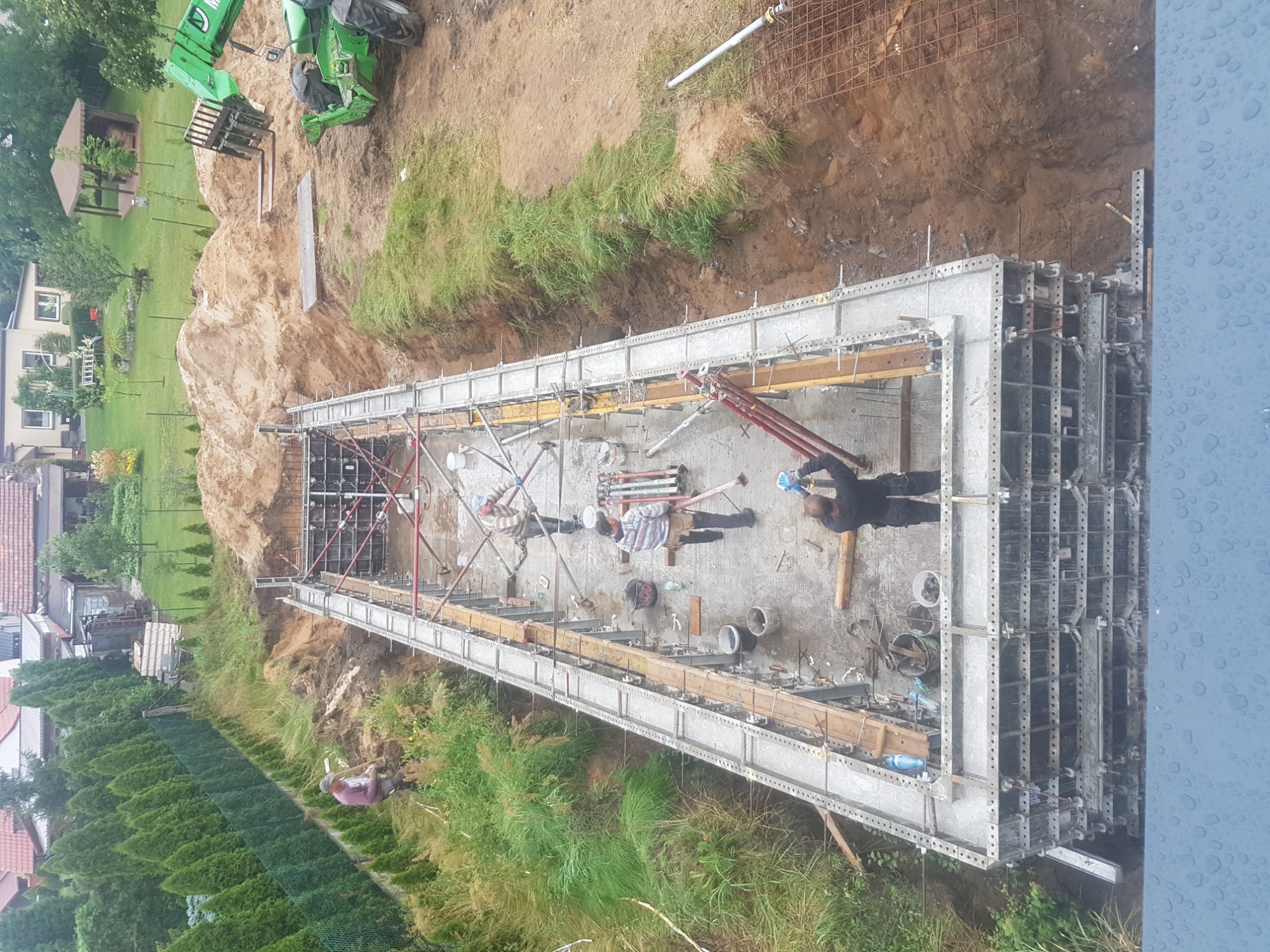 Budowa fundamentów pod budynek: szalunek z metalowych paneli, zbrojenie stalowe, widoczni pracownicy budowlani i sprzęt budowlany na placu budowy.