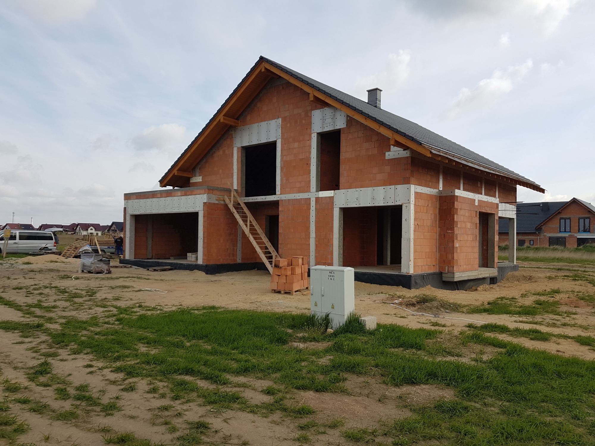 Surowy stan domu jednorodzinnego z czerwonej cegły z drewnianą konstrukcją dachu i schodami wejściowymi, widoczna skrzynka elektryczna na trawniku.