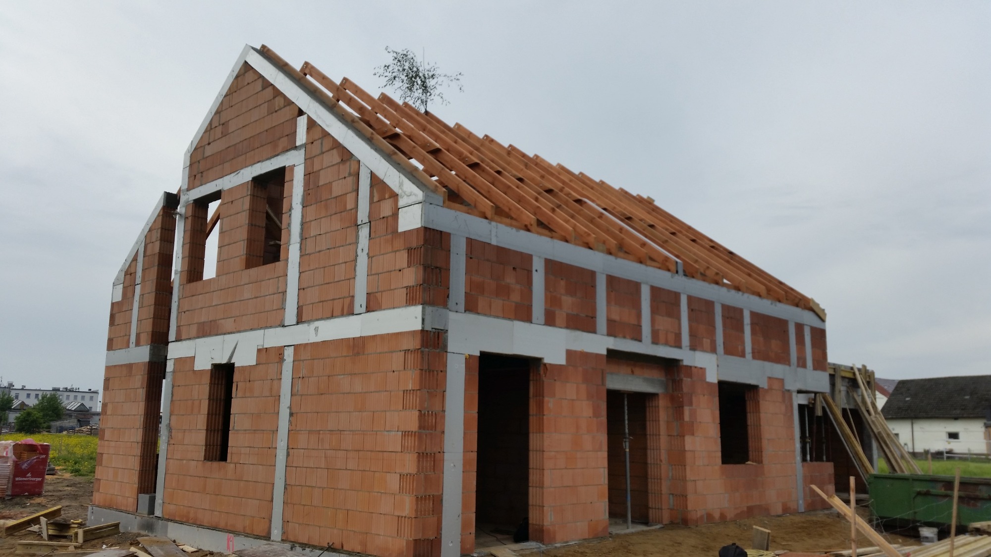 Surowy stan domu murowanego z czerwonej cegły ceramicznej z widoczną konstrukcją dachu i pustymi otworami okiennymi, na tle pochmurnego nieba i zieleni.