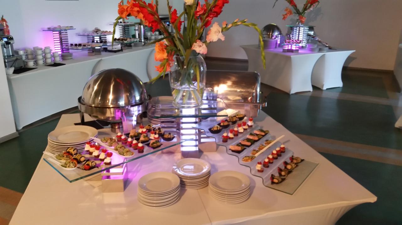 Elegancko przygotowany stół bufetowy z metalowymi podgrzewaczami, szklanymi półmiskami z przekąskami, stosami białych talerzy i dekoracyjnym bukietem z mieczyków w szklanym wazonie, oświetlony...
