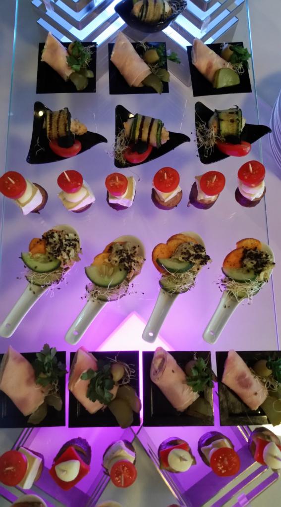 Różnorodne przekąski na szklanym stojaku: roladki z szynki z ziołami, koreczki z pomidorkami i serem, roladki z grillowanej cukinii na czarnych talerzykach, oraz przystawki na białych łyżkach...