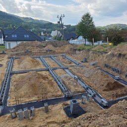 PHU WILBUD Krzysztof Wilczyński - Fundamenty domu jednorodzinnego w trakcie budowy, widok z góry na wykop z izolacją i bloczkami betonowymi, w tle zabudowa mieszkalna i zalesione wzgórza.