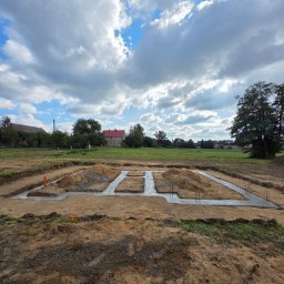 PHU WILBUD Krzysztof Wilczyński - Zalane fundamenty pod budowę domu jednorodzinnego na działce z widokiem na wiejski krajobraz i błękitne niebo z chmurami. Widoczne wystające pręty zbrojeniowe.