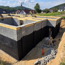 PHU WILBUD Krzysztof Wilczyński - Fundamenty domu z bloczków betonowych, zabezpieczone czarną izolacją, z drewnianym szalunkiem na górze. Widoczne otoczenie budowy i domy w tle.