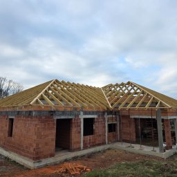 PHU WILBUD Krzysztof Wilczyński - Niski dom w trakcie budowy: czerwona cegła, drewniana więźba dachowa, betonowe słupy. Widok z zewnątrz, pochmurne niebo w tle. Częściowo ułożone cegły na ziemi.