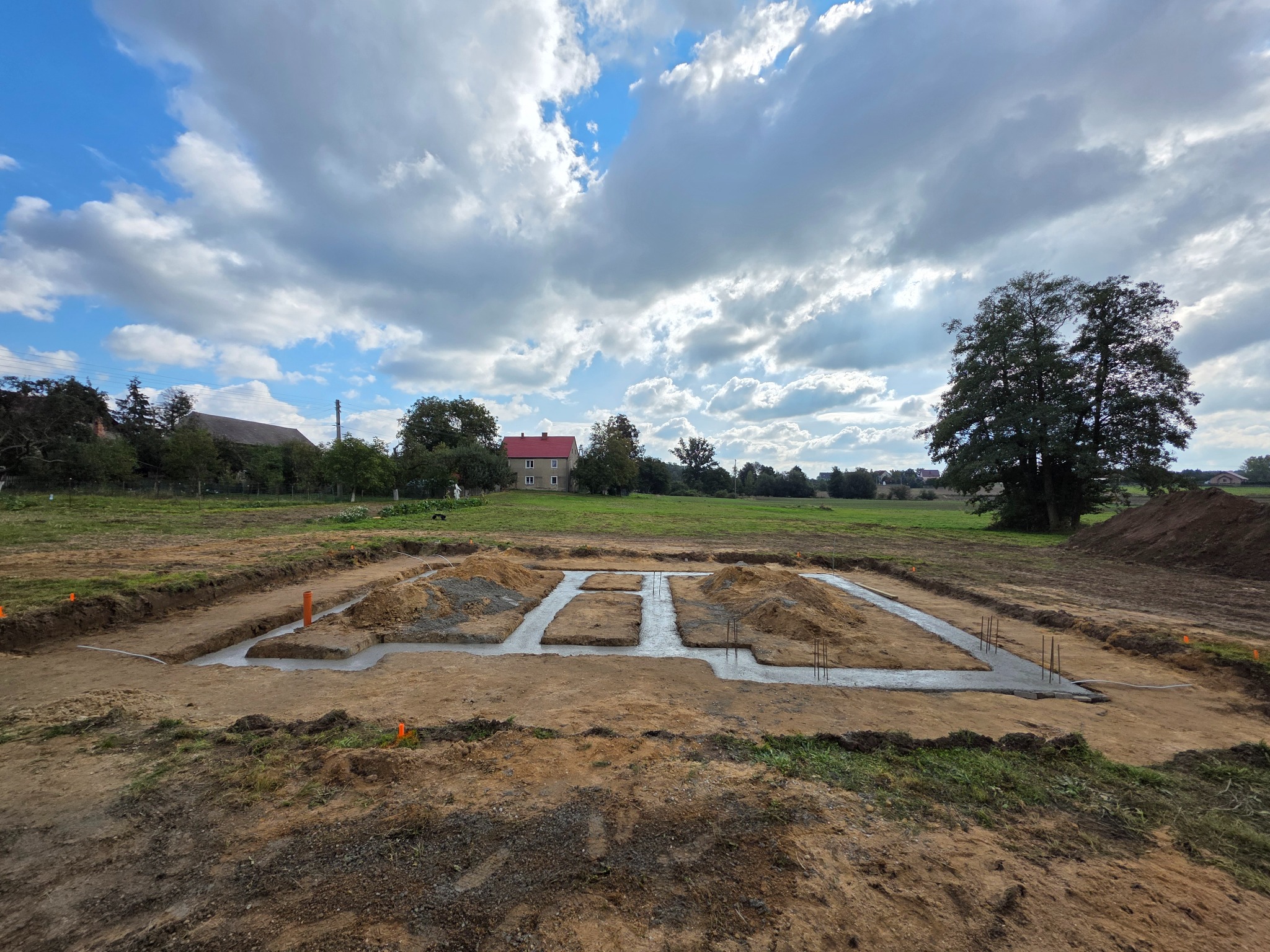 Zalane fundamenty pod budowę domu jednorodzinnego na działce z widokiem na wiejski krajobraz i błękitne niebo z chmurami. Widoczne wystające pręty zbrojeniowe.