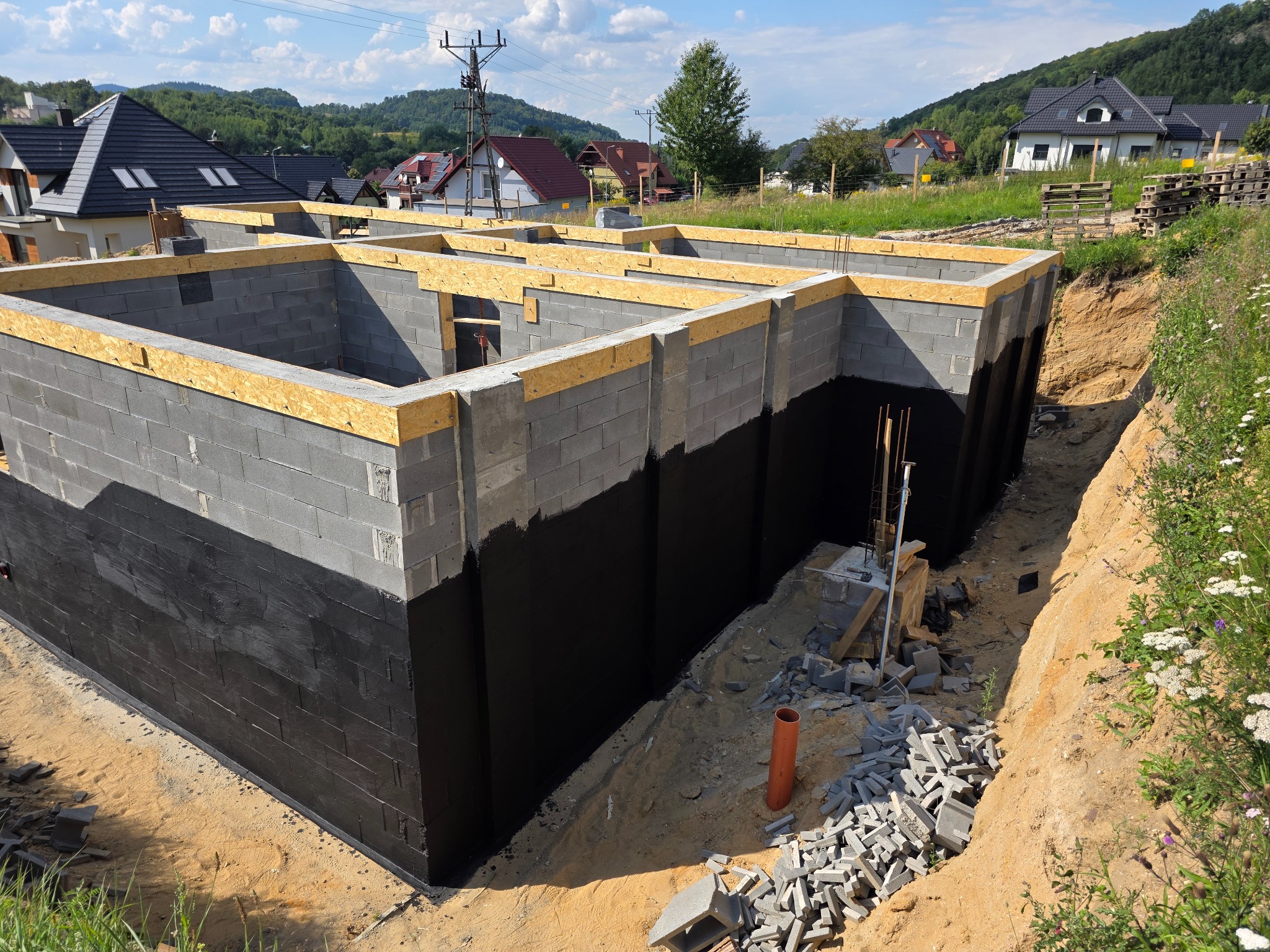 Fundamenty domu z bloczków betonowych, zabezpieczone czarną izolacją, z drewnianym szalunkiem na górze. Widoczne otoczenie budowy i domy w tle.