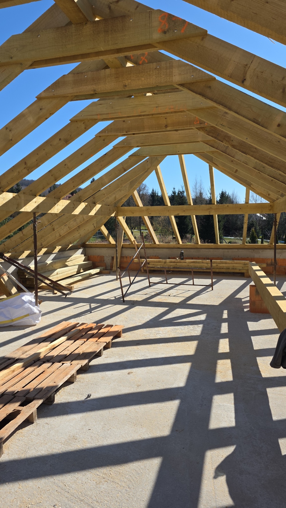 Drewniana konstrukcja więźby dachowej w trakcie montażu, widok z wnętrza budynku, jasne niebo w tle. Elementy konstrukcyjne z widocznymi oznaczeniami.