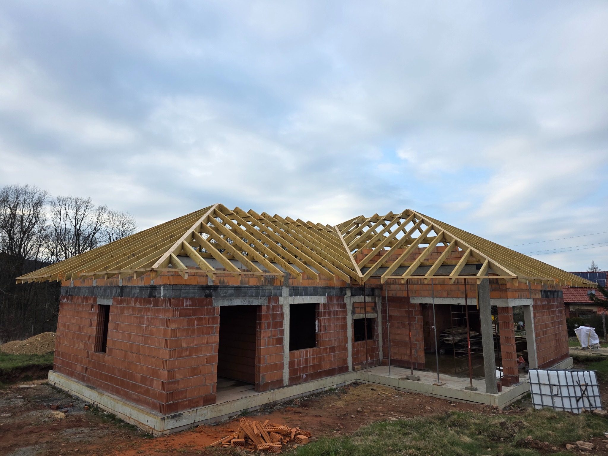 Niski dom w trakcie budowy: czerwona cegła, drewniana więźba dachowa, betonowe słupy. Widok z zewnątrz, pochmurne niebo w tle. Częściowo ułożone cegły na ziemi.