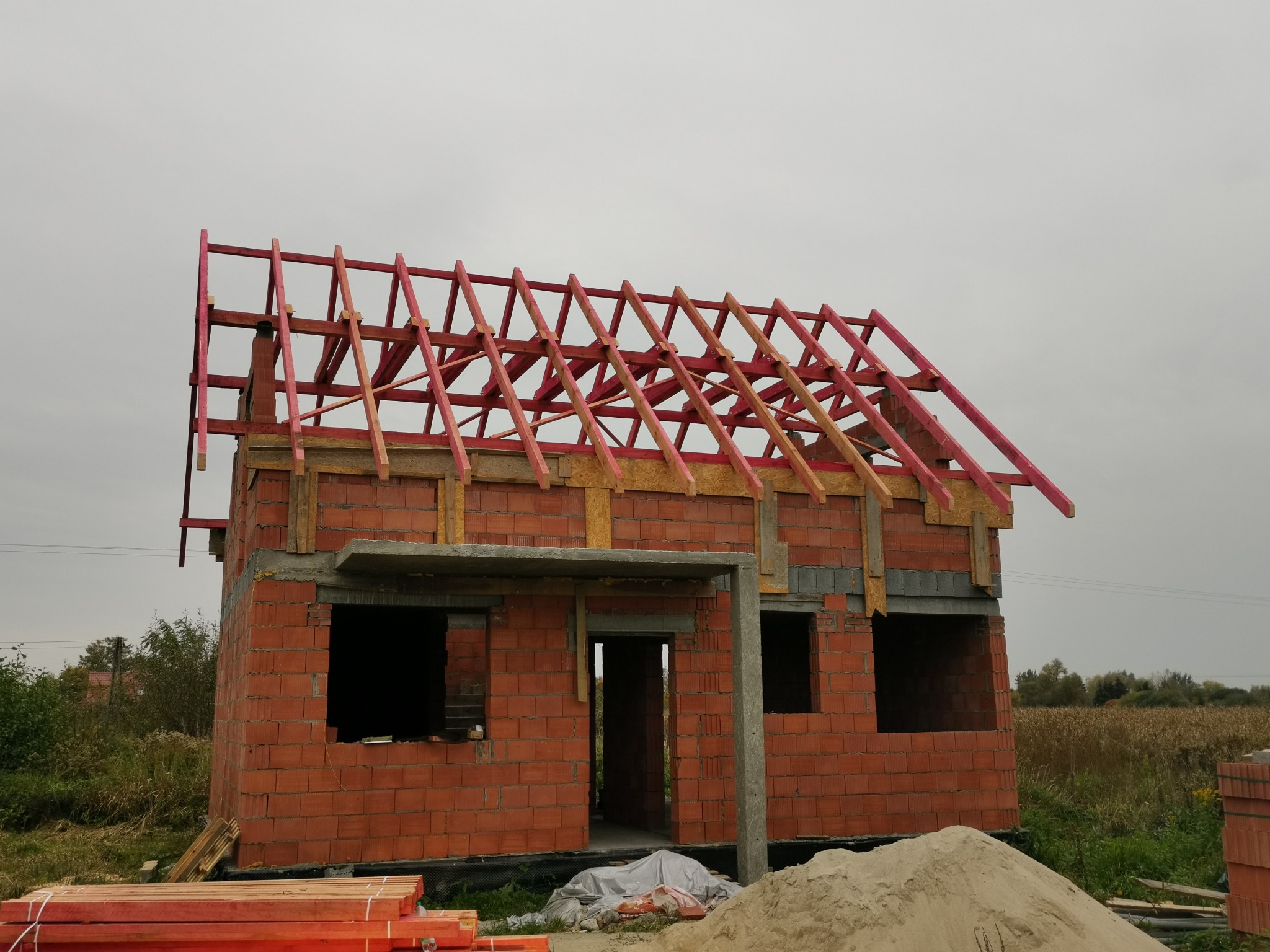 Niedokończony dom z czerwonej cegły z drewnianą konstrukcją dachu pomalowaną na różowo, na tle pochmurnego nieba i pola. Widoczne materiały budowlane.