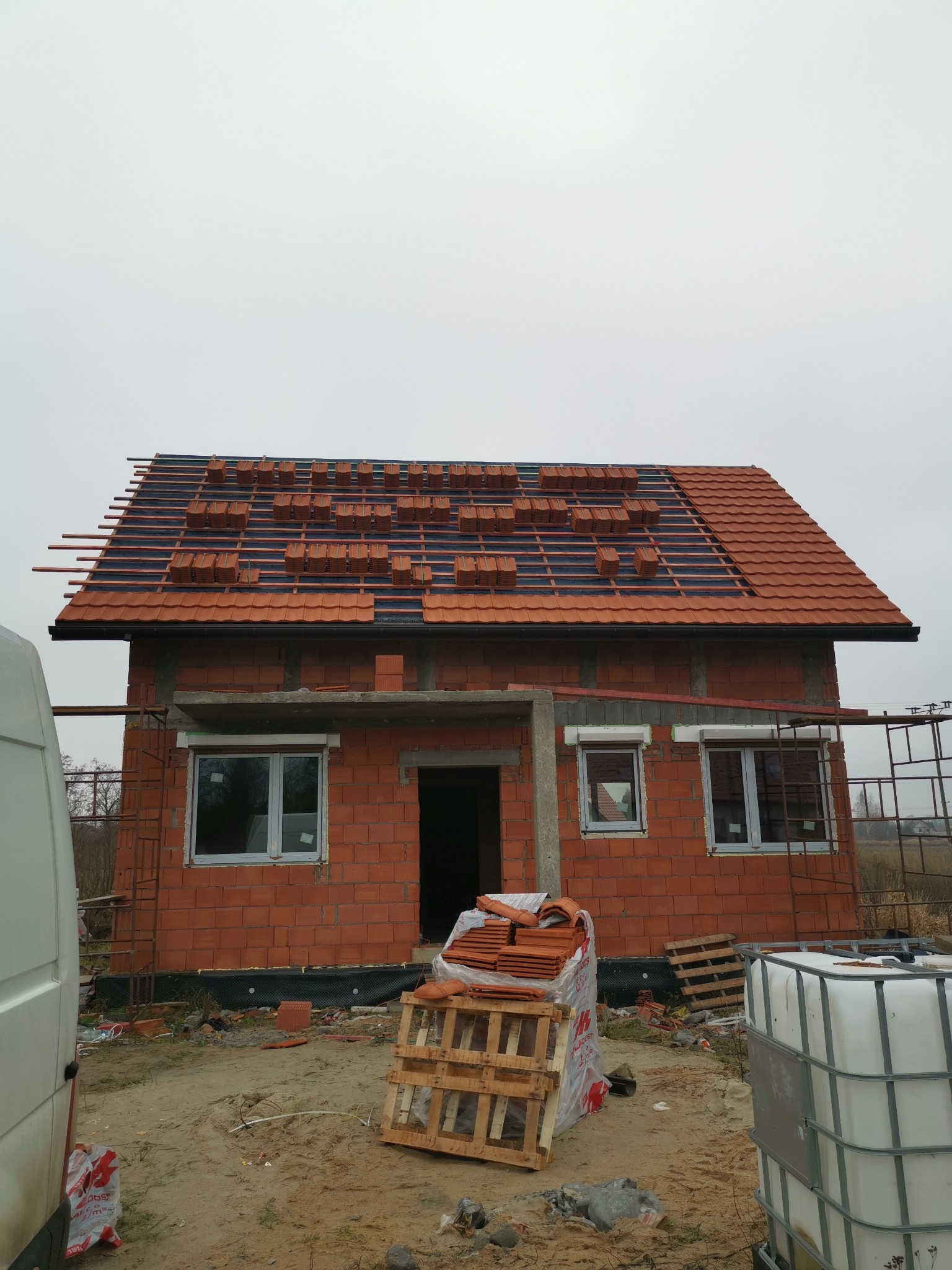 Budowa domu z czerwonej cegły: częściowo ułożona dachówka na łatach, okna wstawione, paleta z dachówkami na ziemi. Widok na budynek w trakcie prac dekarskich.