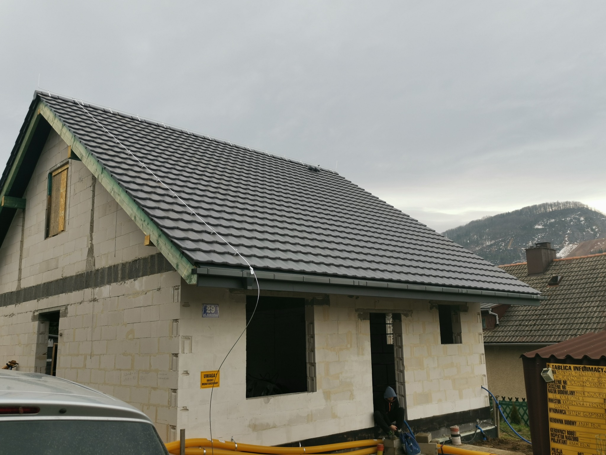 Dom w trakcie budowy z szarym dachem z dachówki, widoczne bloczki betonu komórkowego i częściowo zamurowane okna. Na ścianie tabliczka z numerem posesji.
