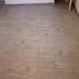 Ułożona podłoga z szarych paneli imitujących drewno, widoczne fugi i krawędzie ścian, jasne tło.