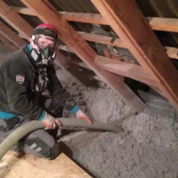 Pracownik w masce ochronnej aplikuje szarą izolację celulozową w przestrzeni poddasza za pomocą węża, widoczne drewniane krokwie i deski szalunkowe.