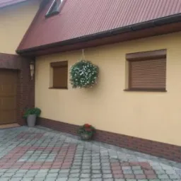 Dom jednorodzinny z brązowymi roletami zewnętrznymi, wiszącą kulą kwiatową i numerem domu 69.