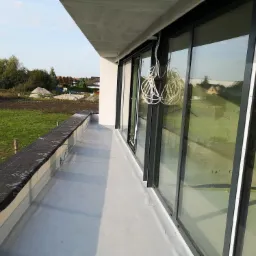 Wykończony płaski dach z papy termozgrzewalnej na balkonie nowoczesnego budynku z dużymi przesuwnymi oknami, widoczne przewody elektryczne pod sufitem.