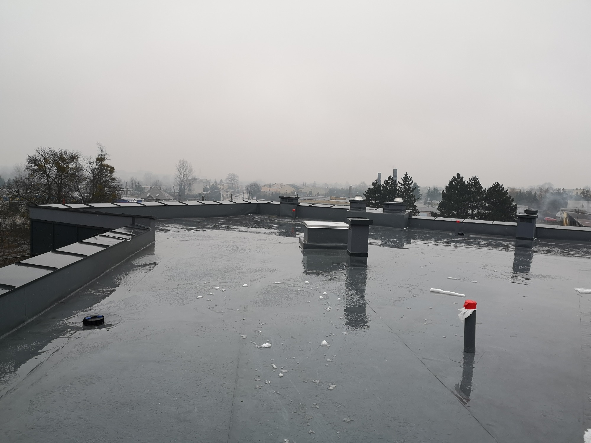 Szary płaski dach pokryty papą termozgrzewalną w pochmurny dzień, widoczne kominy wentylacyjne i obróbki blacharskie na krawędzi dachu, w tle zabudowa miejska i drzewa.
