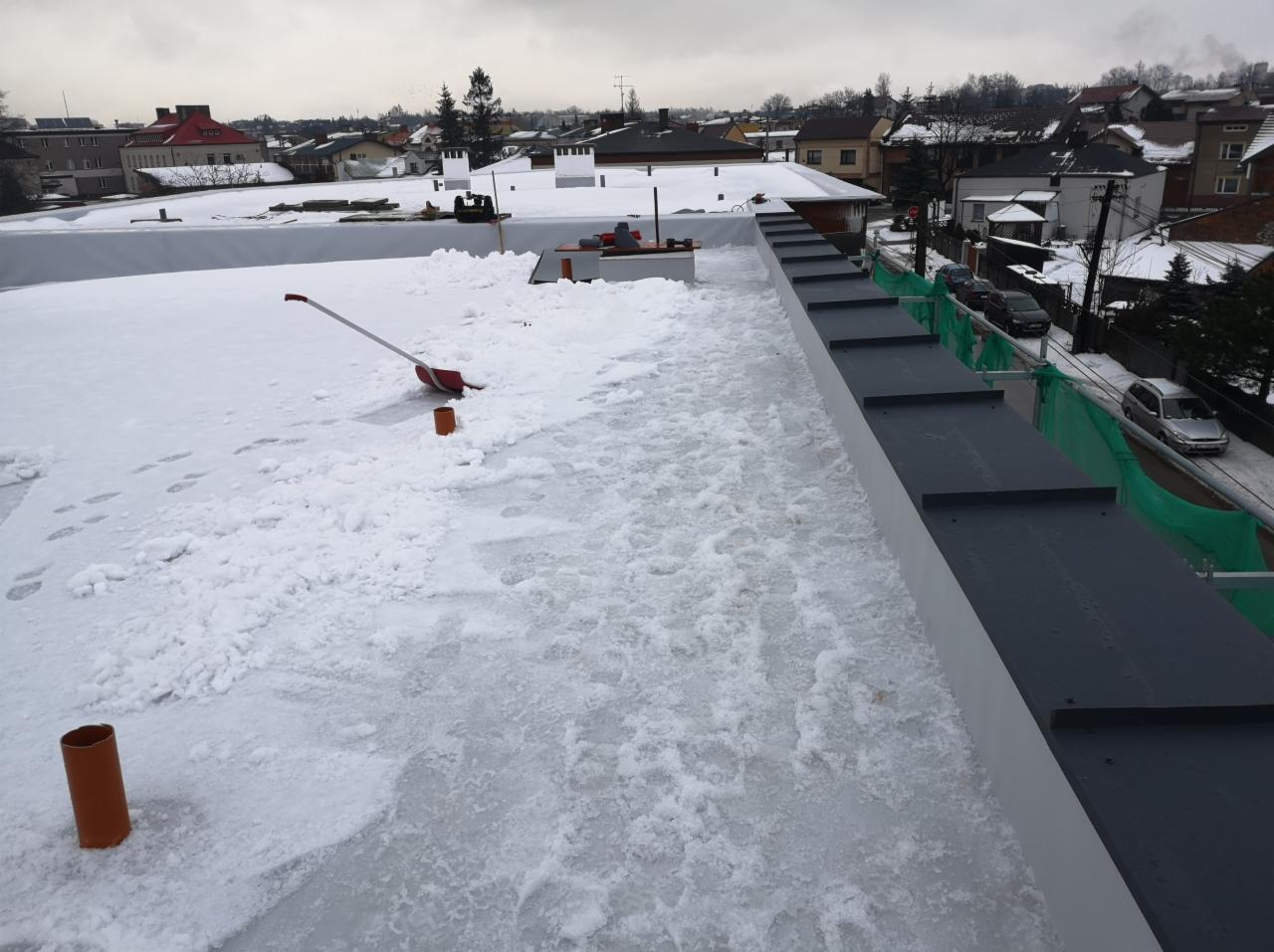 Płaski dach pokryty śniegiem i lodem, częściowo odśnieżony, widoczne ślady stóp, łopata do śniegu i fragment obróbki blacharskiej w tle zabudowa miejska.
