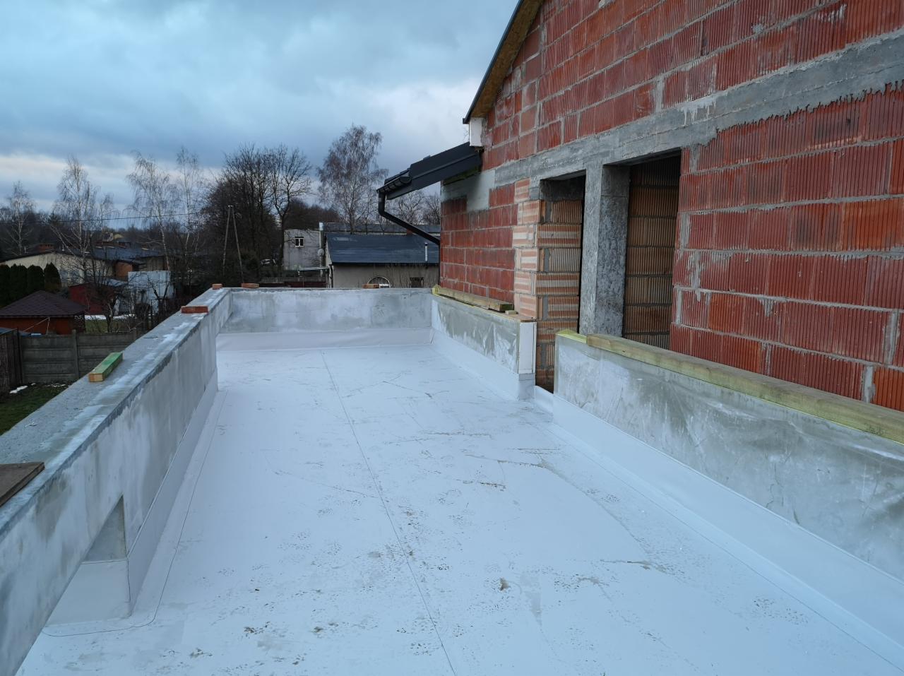 Biała, płaska powierzchnia dachu z wykończonymi krawędziami, w tle budynek w trakcie budowy z czerwonej cegły i ponure niebo.