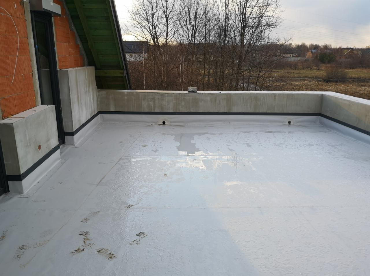 Płaski dach pokryty białą, płynną membraną hydroizolacyjną z widocznymi wpustami dachowymi i fragmentami muru budynku w tle.