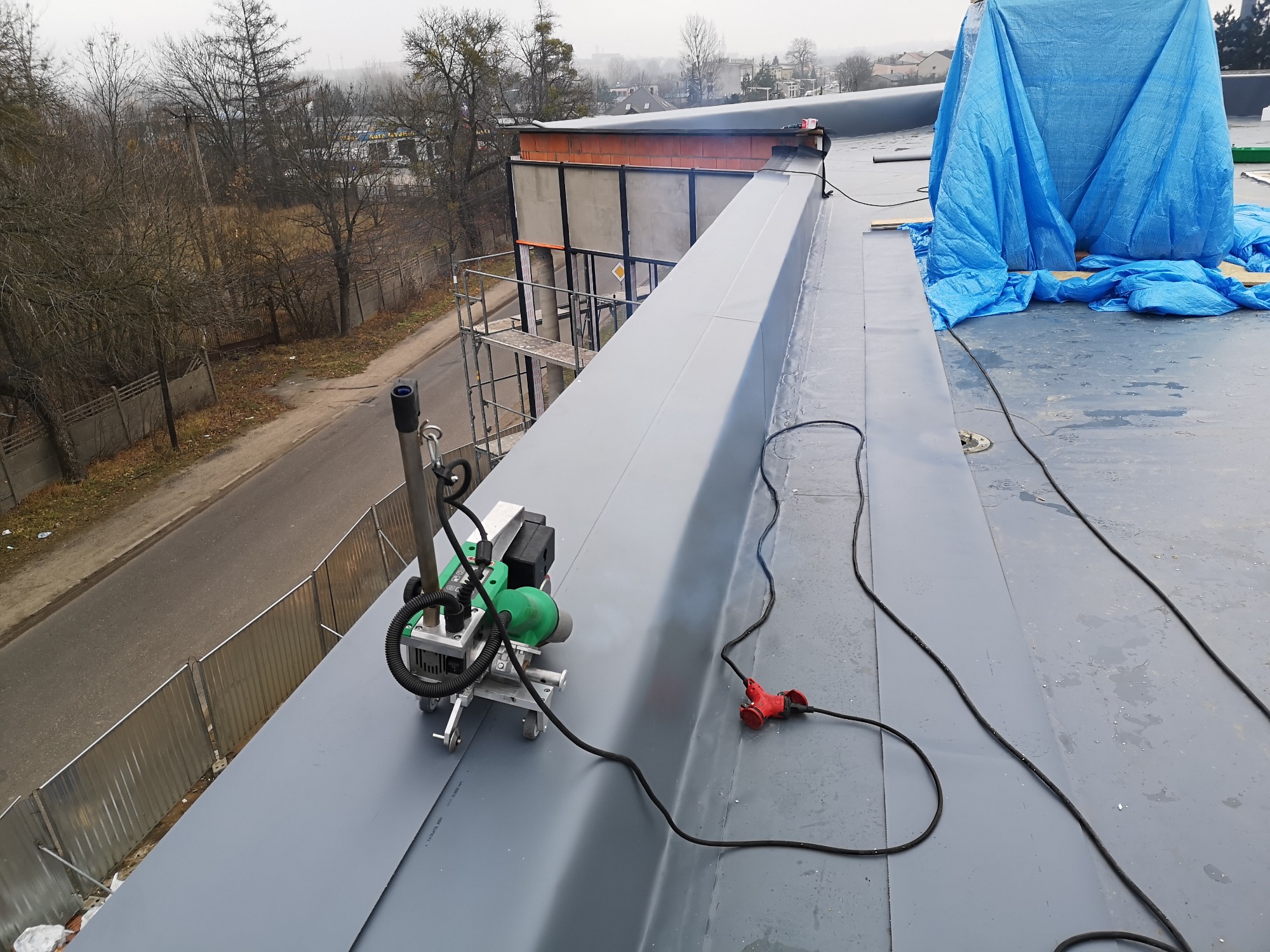 Szary dach płaski z urządzeniem do zgrzewania membrany dachowej, widoczne kable, w tle budynek w trakcie budowy i drzewa.