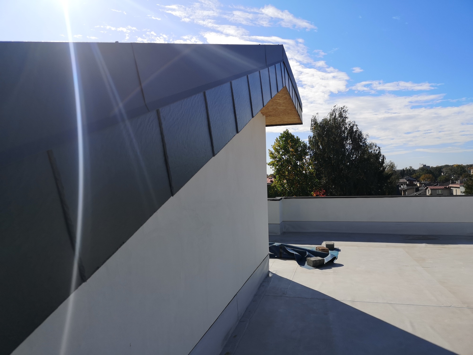 Nowoczesna, czarna blacha na dachu budynku z płaskim dachem, widok z boku, na dachu leżą materiały budowlane, w tle błękitne niebo z chmurami i zieleń drzew.