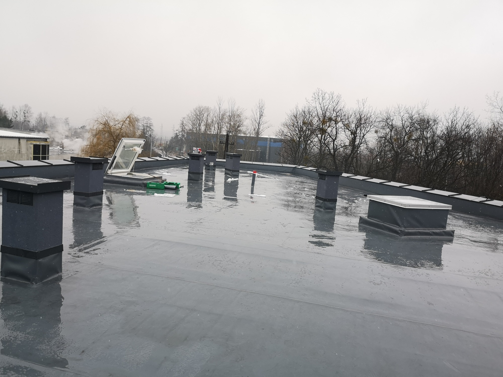 Szary, płaski dach pokryty papą z wywietrznikami i świetlikiem dachowym w pochmurny dzień.