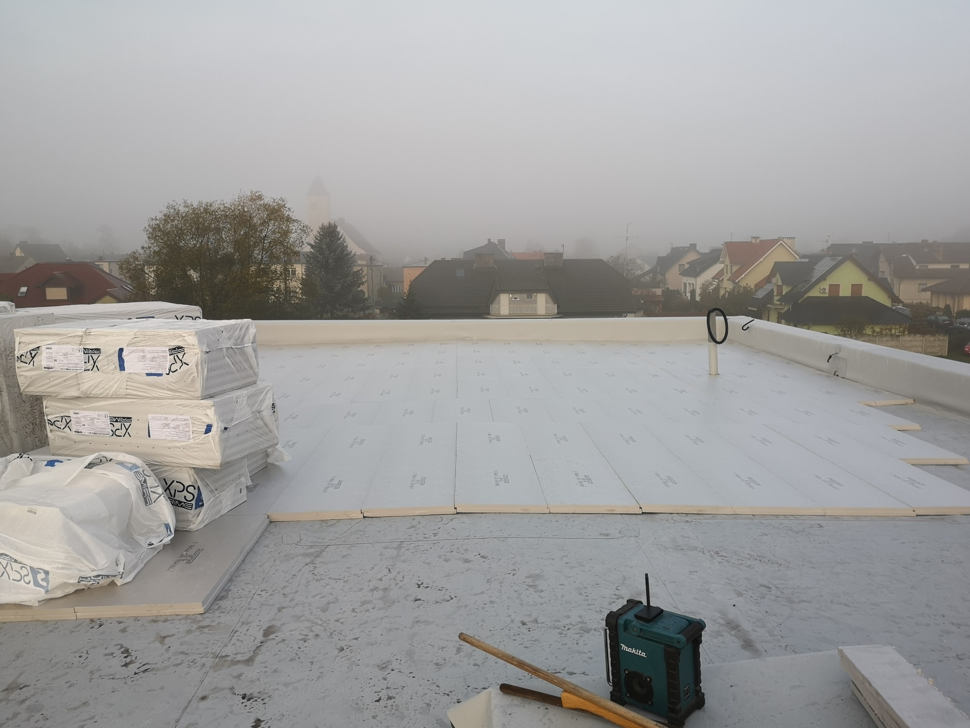 Płaski dach pokryty białymi płytami izolacyjnymi, z zapasem materiałów i radiem budowlanym na pierwszym planie, w tle zamglony krajobraz miejski.