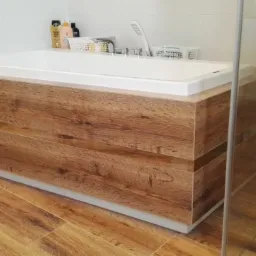 Nowoczesna obudowa wanny z drewnopodobnych paneli, dopasowana do podłogi, biała wanna z armaturą i akcesoriami w tle, kabina prysznicowa z przezroczystego szkła.