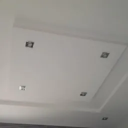 Biały sufit podwieszany z czterema kwadratowymi lampami halogenowymi, widoczne fragmenty ściany z płytkami.