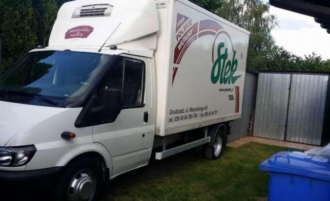 Biały samochód dostawczy z chłodnią, oznaczony logo i danymi kontaktowymi firmy 'Staro Polskie Wędliny', zaparkowany obok garażu i niebieskiego pojemnika na śmieci.