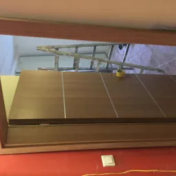 Widok przez ościeżnicę na fragment ściany z żółtą taśmą malarską i drabiną w tle, czerwona ściana poniżej ościeżnicy z widocznym gniazdkiem elektrycznym.