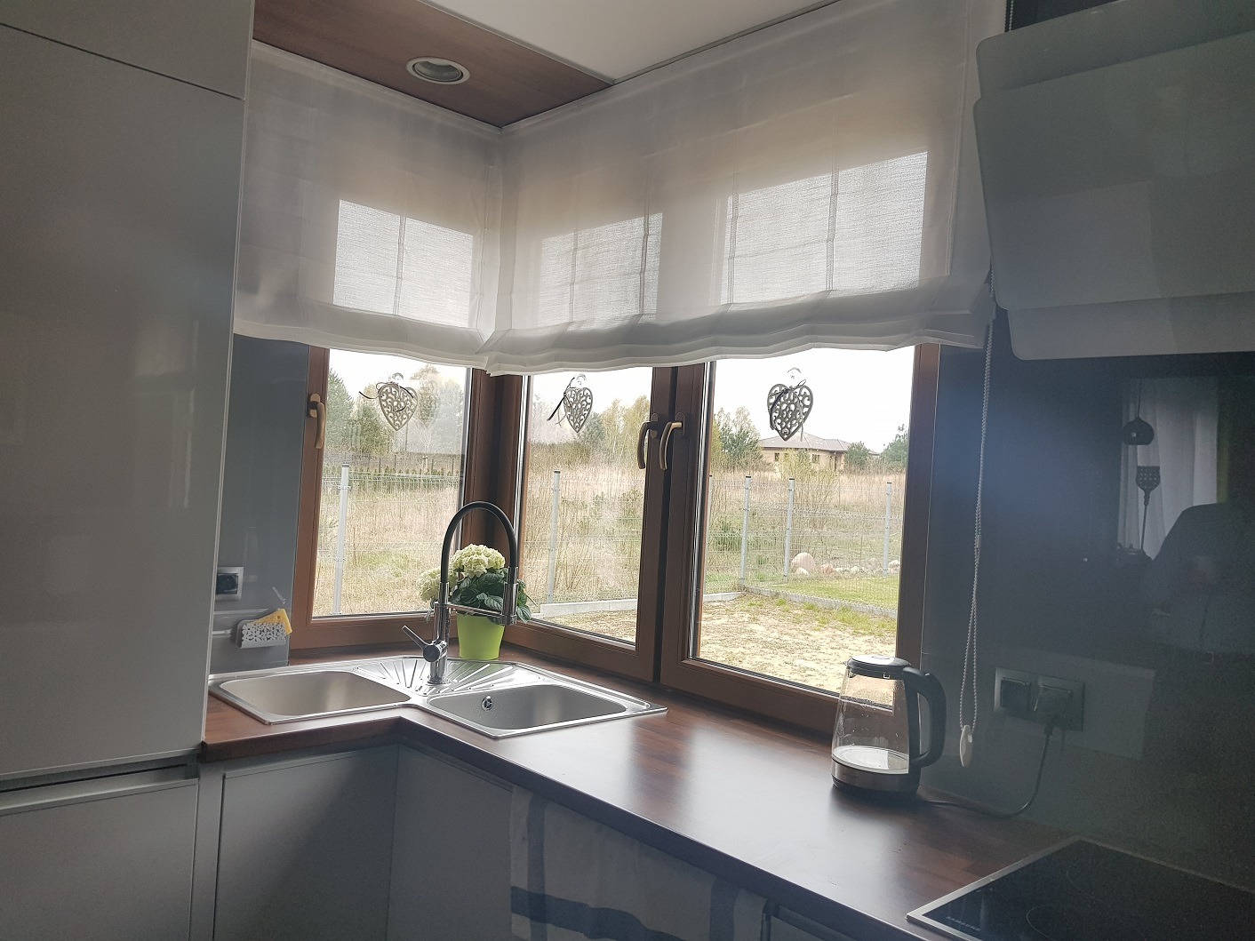 Jasne, zwiewne rolety rzymskie w kuchni w Luboniu. Delikatna tkanina, podciągnięta do góry, odsłania widok na zewnątrz. Okna z dekoracyjnymi zawieszkami w kształcie serc.