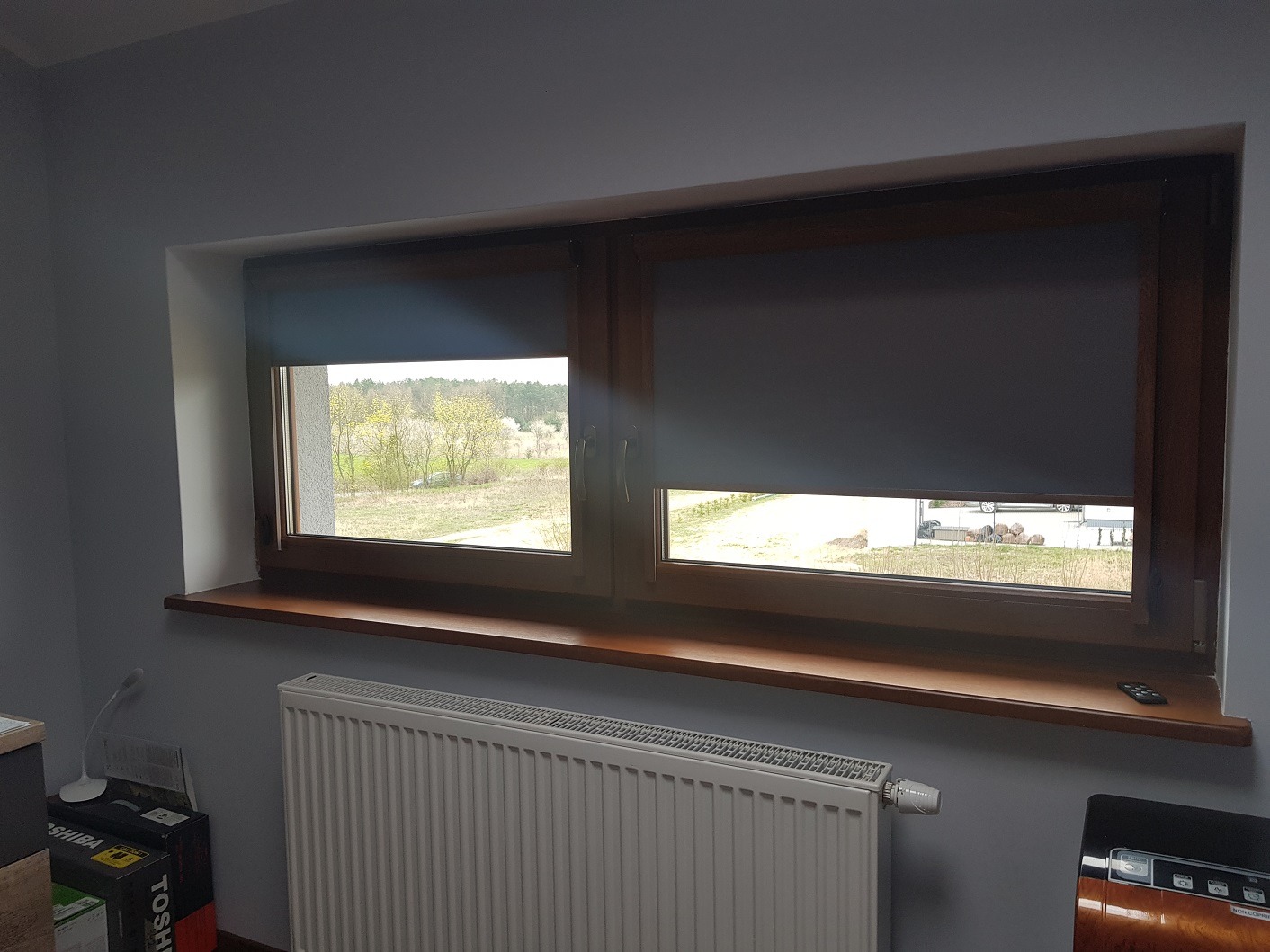 Dwa okna z drewnianą ramą, każde z szarą roletą. Jedna roleta jest częściowo opuszczona, odsłaniając widok na zewnątrz. Na parapecie leży pilot.