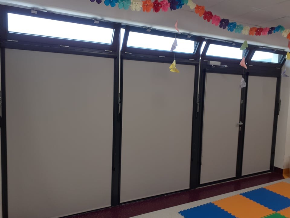 Zasłonięte okna i drzwi z zamontowanymi, szarymi roletami w pomieszczeniu z kolorową dekoracją i podłogą z puzzli piankowych. Widok na całą ścianę.