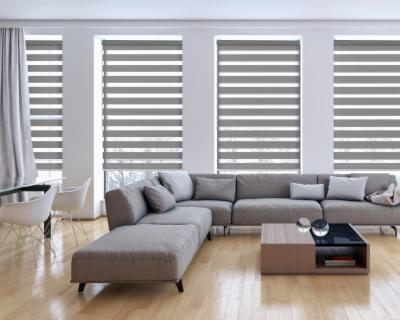 Jasne wnętrze salonu z szarym narożnikiem i trzema oknami przesłoniętymi szarymi roletami w systemie dzień i noc, ustawionymi w pozycji częściowego zaciemnienia.