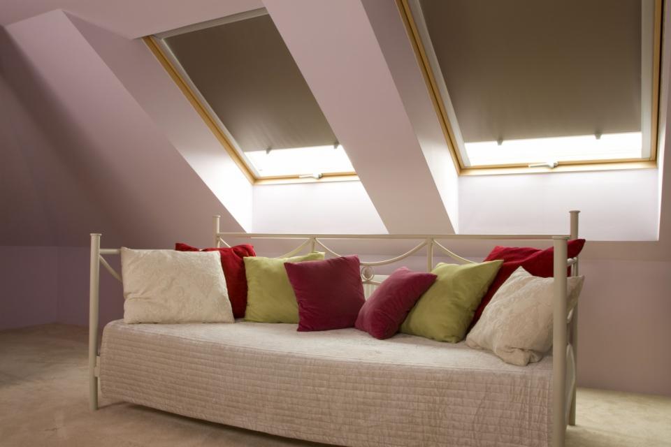 Poddasze z dwoma oknami dachowymi wyposażonymi w szare rolety, pod oknami biała kanapa z kolorowymi poduszkami.
